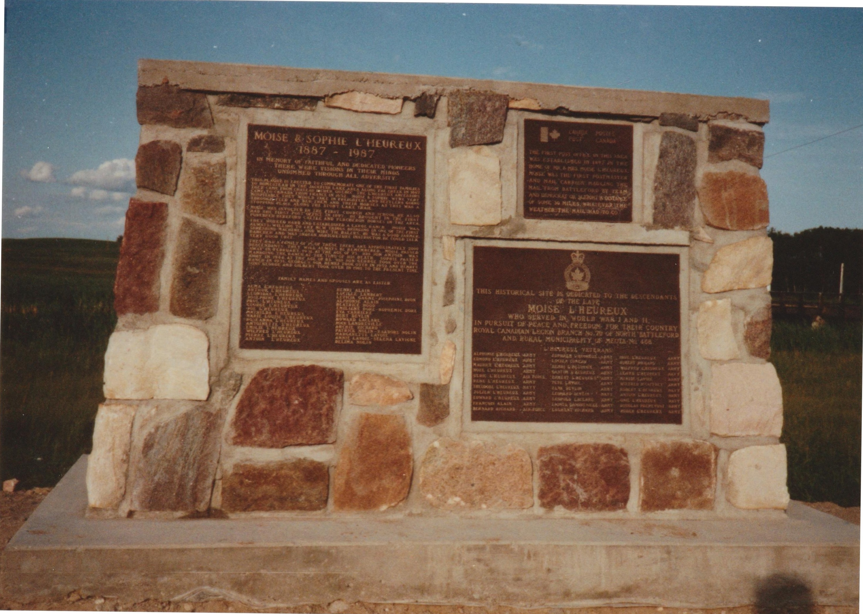 A stone monument with three bronze plaques set into it, standing in a field under a blue sky.